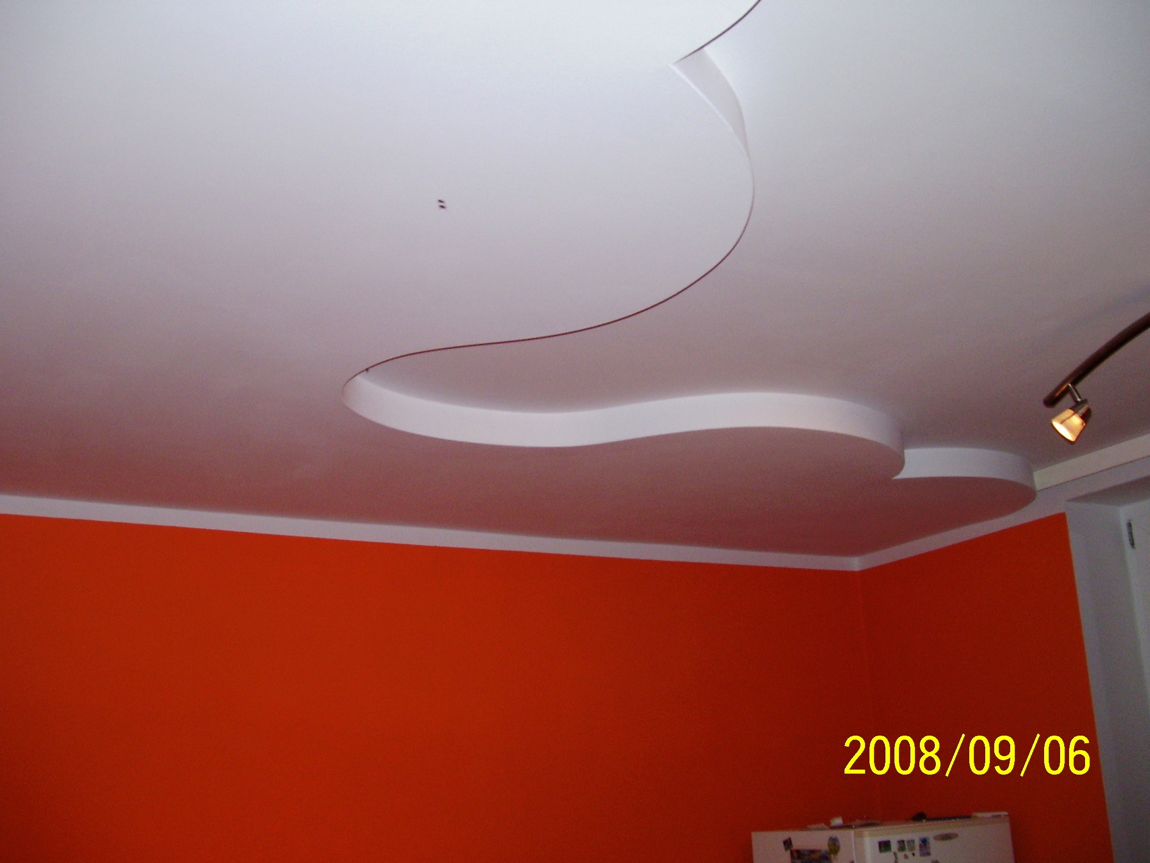 Nowoczesny sufit podwieszany z dekoracyjnym wycięciem w kształcie fali, oświetlenie punktowe i pomarańczowa ściana w tle.